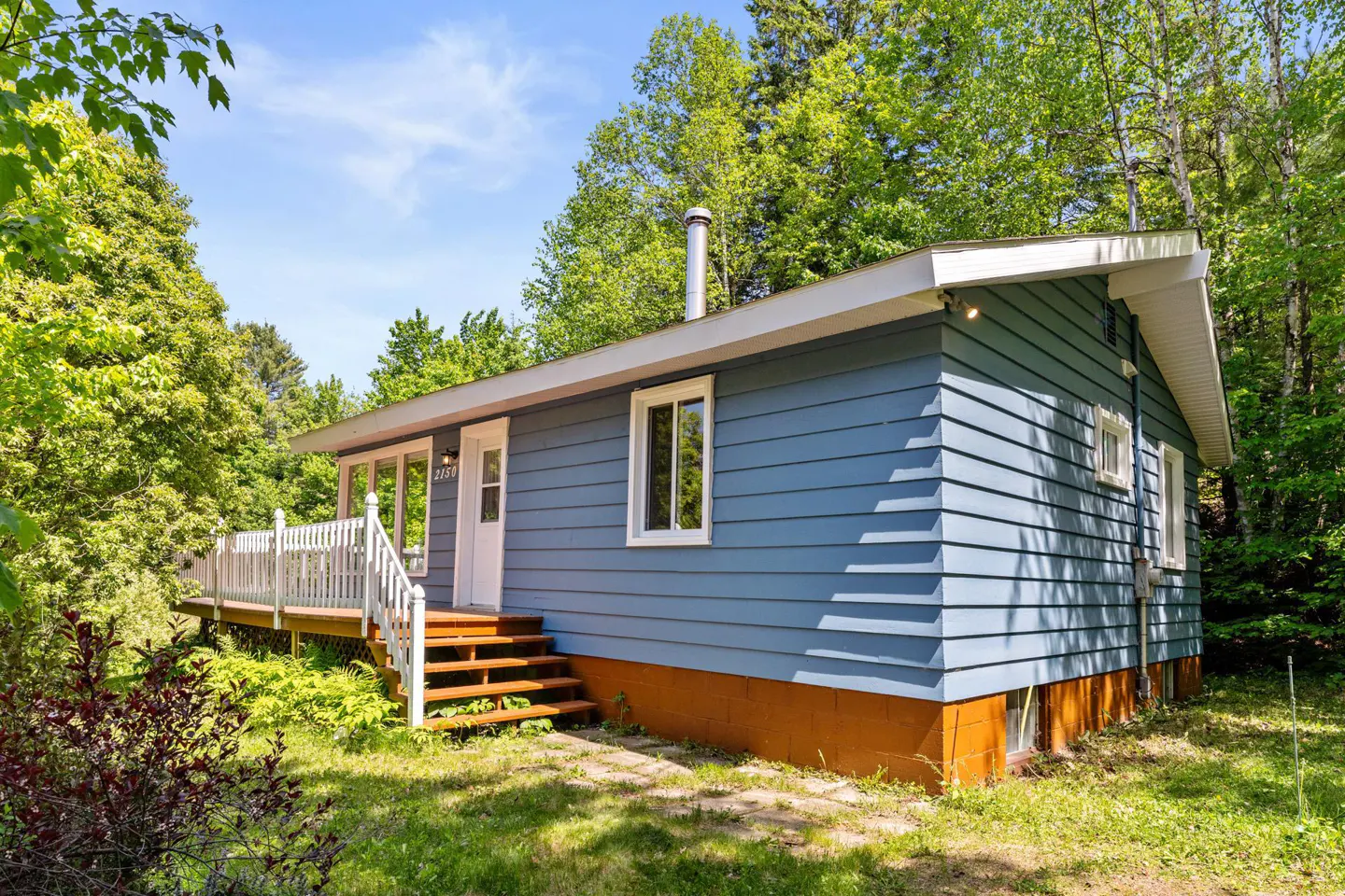 Blue cottage with white trim, a wooden deck, and a brown foundation, surrounded by lush green trees on a sunny day.