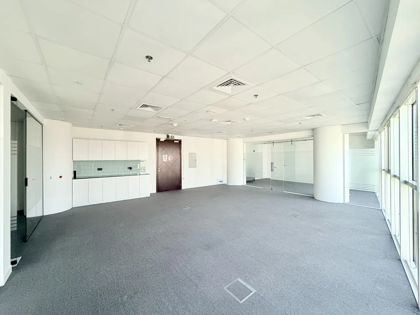 Bright, empty office space with gray carpet, white walls, and large windows. A kitchenette and glass-walled rooms are visible.