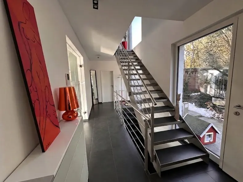Modern home interior with a metal staircase, white walls, and dark tile floors. A red painting and lamp sit on a white dresser. Outside, a small red dog house is visible.