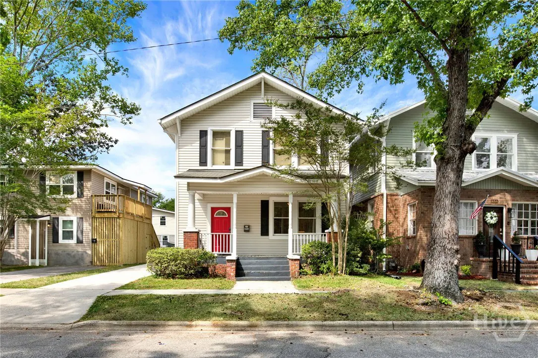 Two-story house with light gray siding, black shutters, and a bright red front door. Green trees frame the house against a blue sky.