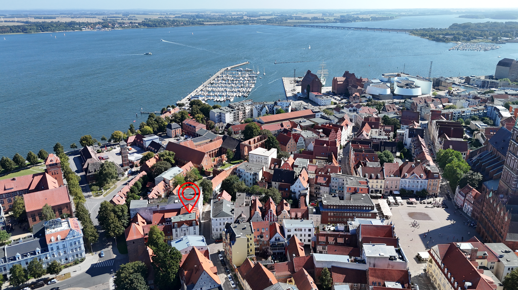 Aerial view of a coastal town with a red location pin marking a specific building near the harbor and city square. [AI-generated text]