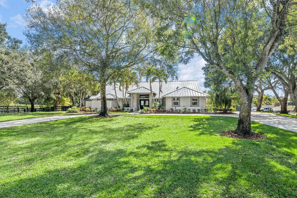 A white, single-story house with a metal roof sits on a large green lawn, framed by mature trees under a blue sky.