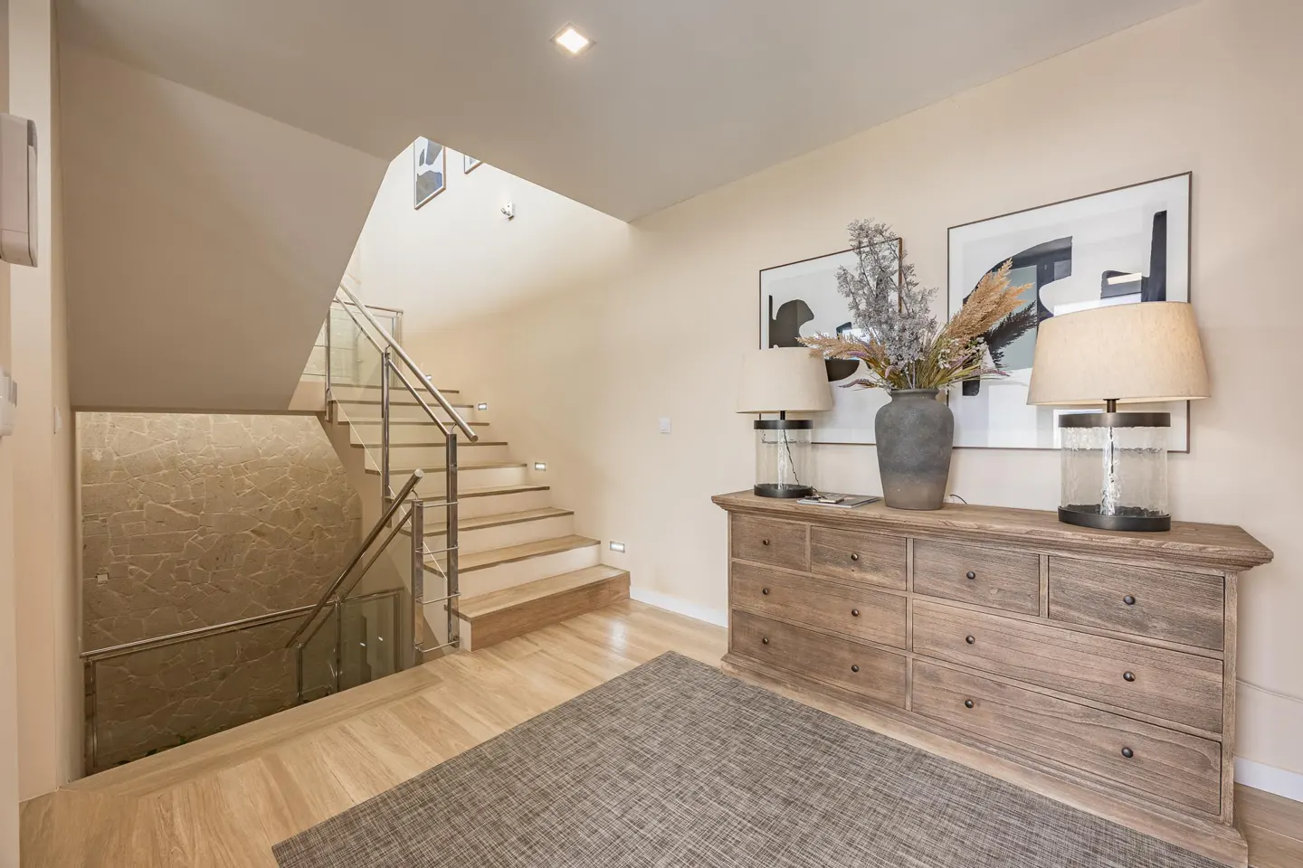 Entryway with a wooden dresser, lamps, and artwork. A staircase with glass railings leads upstairs. A gray rug sits on the wooden floor.