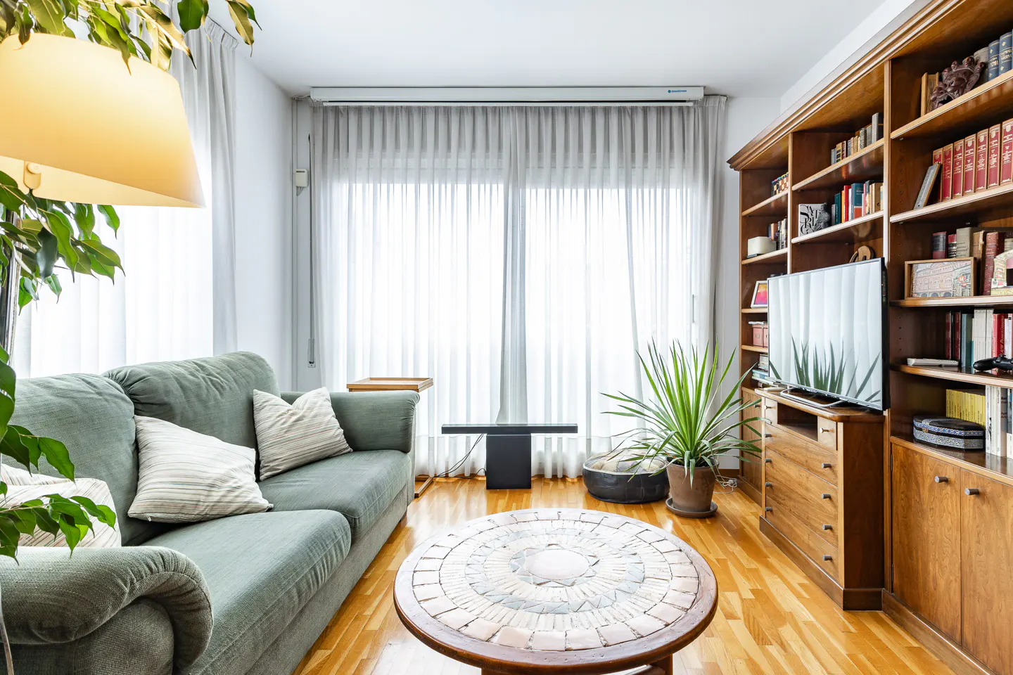 Cozy living room with a green sofa, round mosaic coffee table, large wooden bookshelf, and sheer curtains over a window.