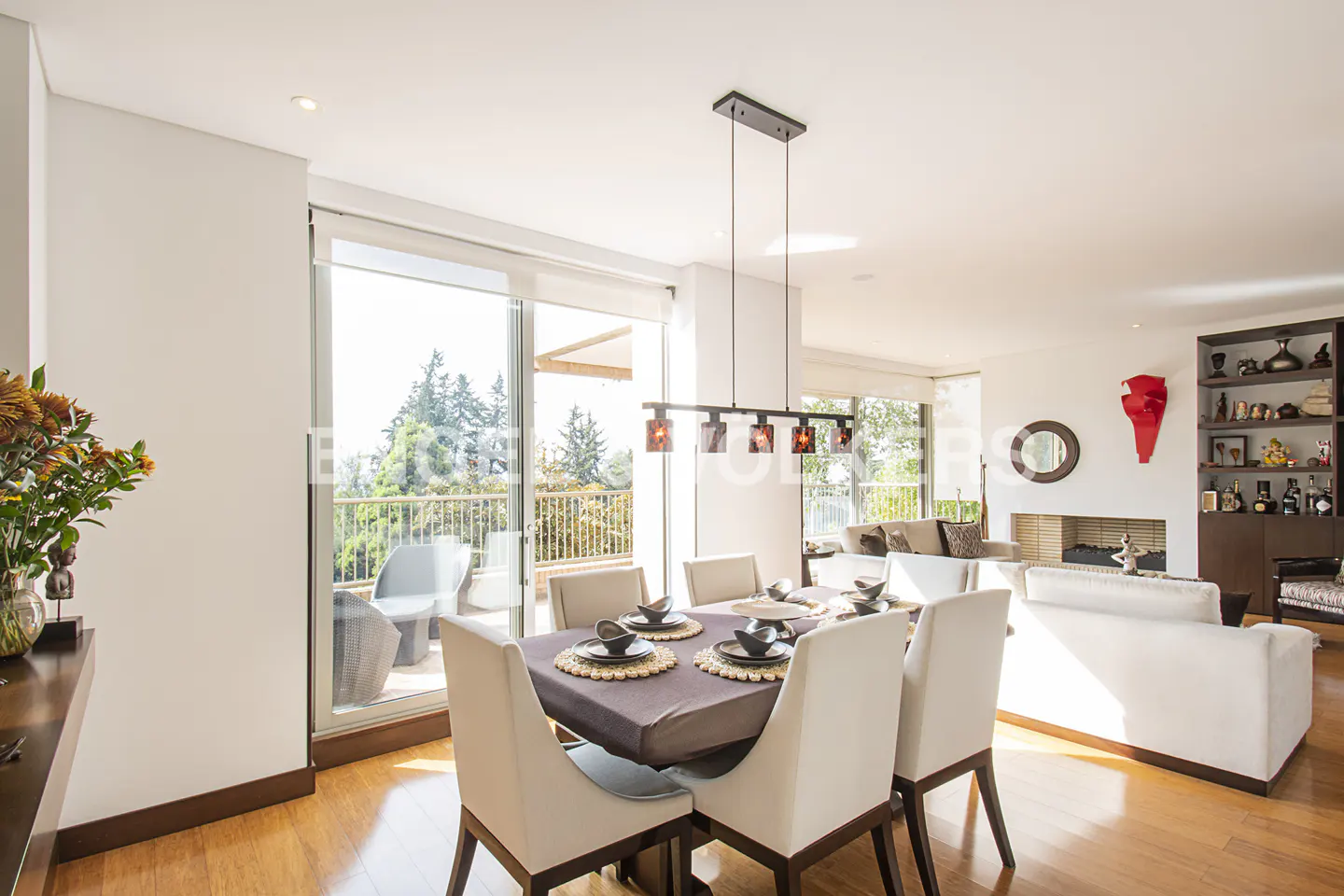 Bright, open-plan dining and living area with wood floors. A table with six chairs is set for a meal. Balcony with trees visible.