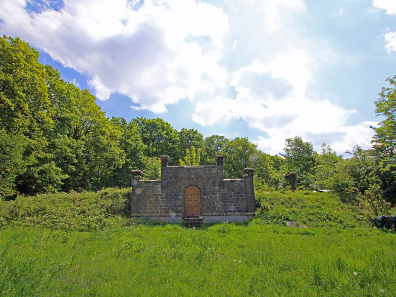 A stone structure with a wooden door set into a grassy hillside, surrounded by lush green trees under a cloudy blue sky.