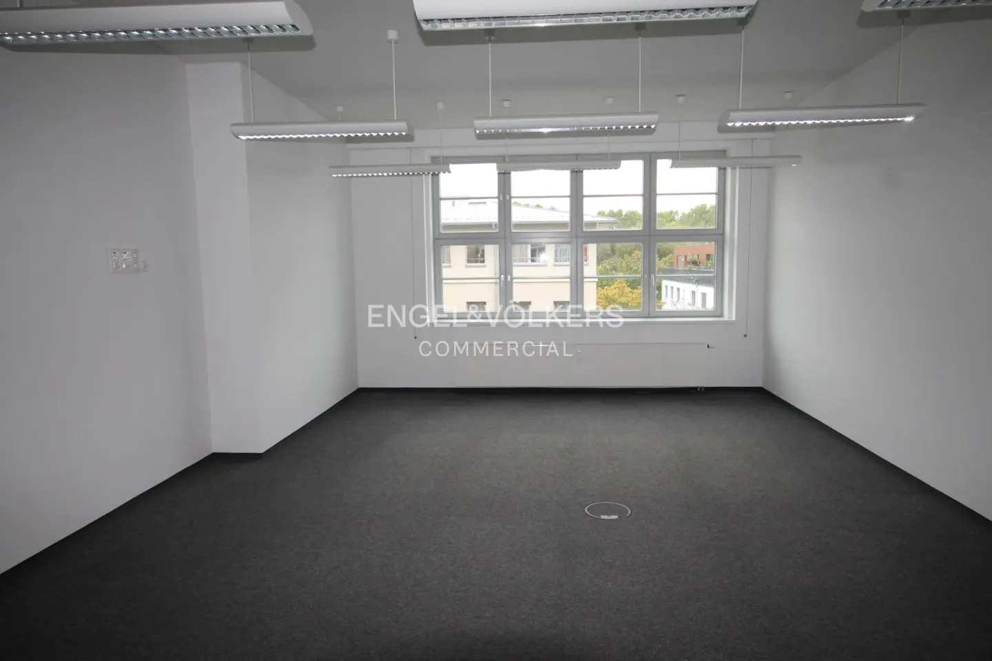 Empty office space with gray carpet, white walls, and a large window showing a building and trees. Overhead lighting fixtures are visible.