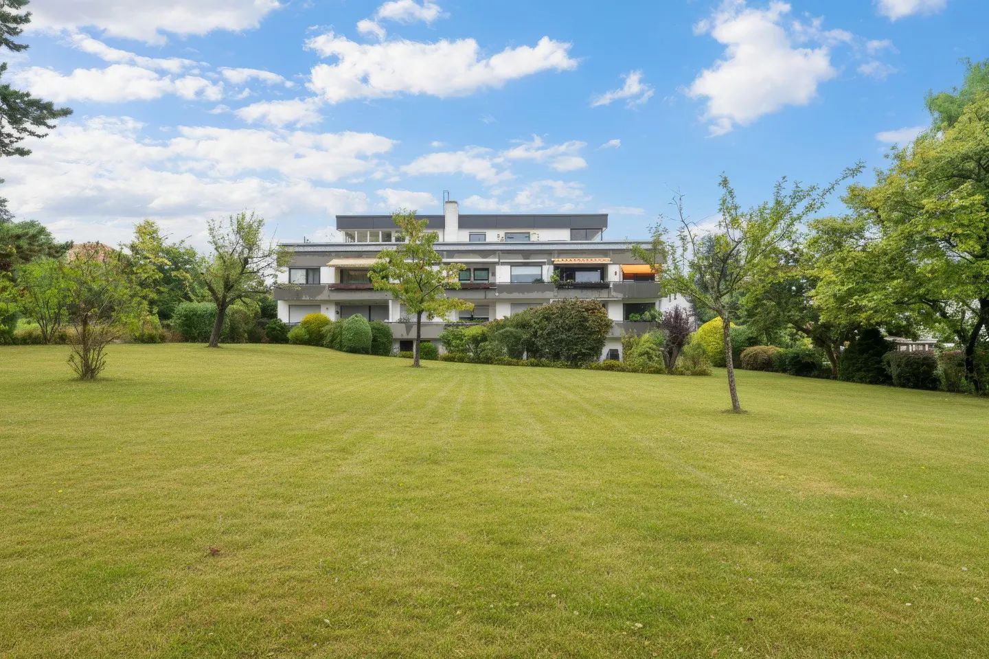 A three-story white apartment building with balconies, surrounded by a green lawn and trees under a blue sky.