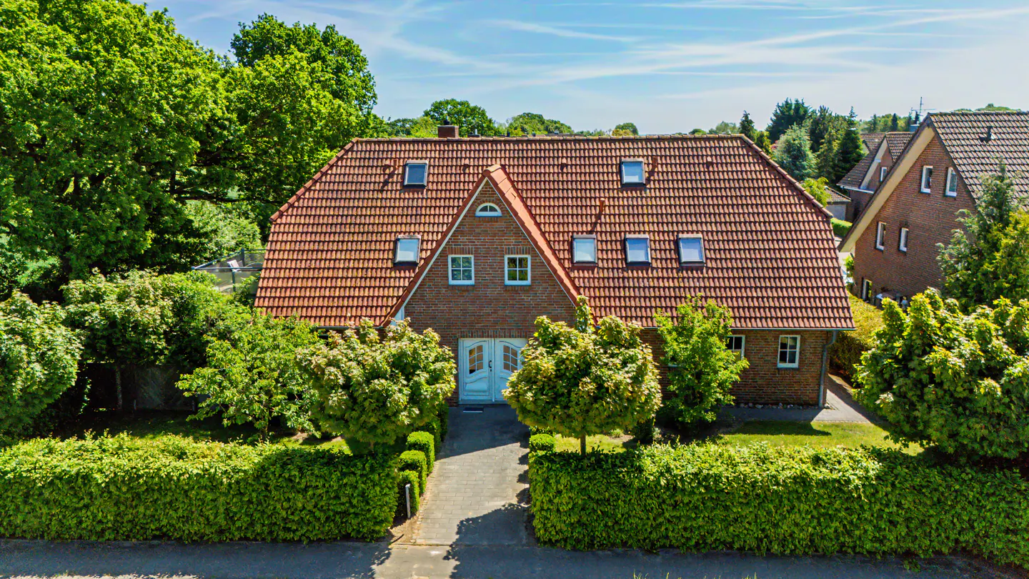 A two-story brick house with a red tile roof, white front doors, and multiple skylights, surrounded by lush green hedges.