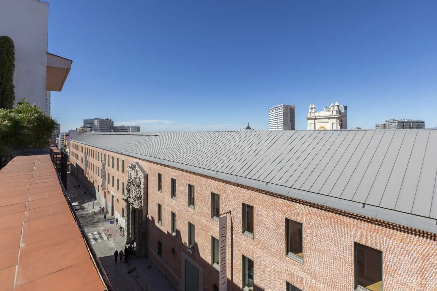 High-angle view of the red brick Conde Duque building in Madrid with a long metal roof under a clear blue sky.