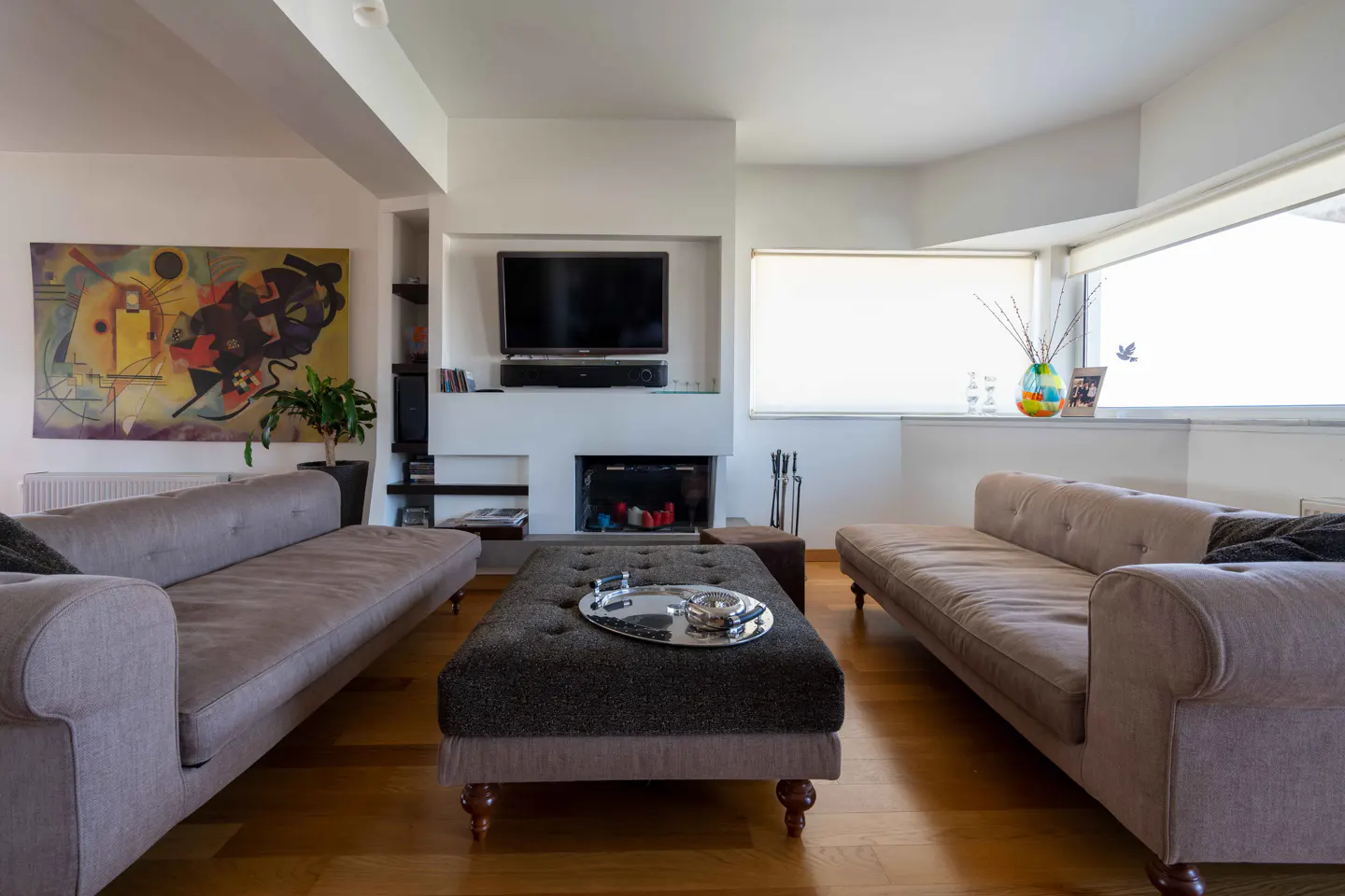 Living room with two beige sofas facing each other, a dark ottoman, and a fireplace with a TV above it. A colorful painting hangs on the wall.
