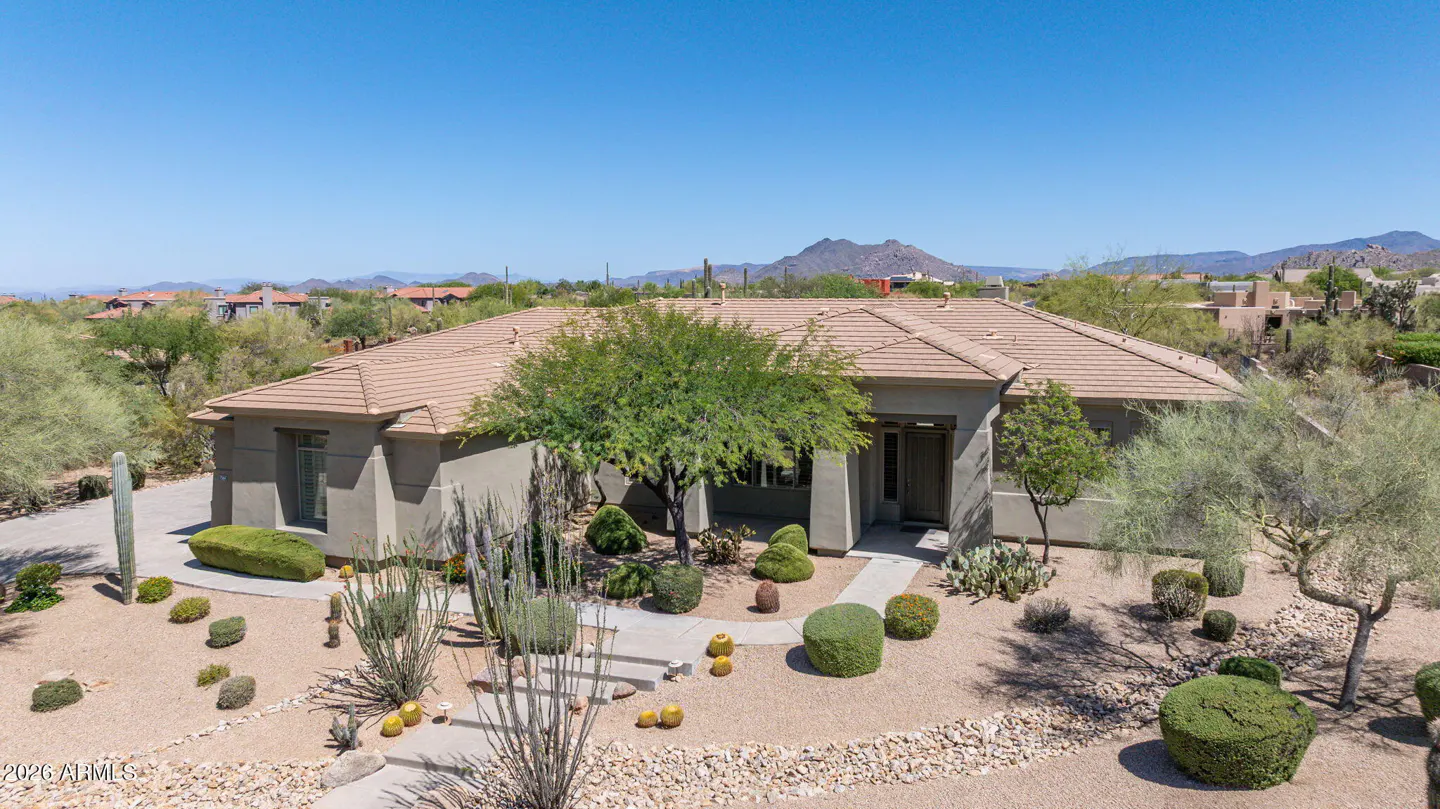 Aerial view of a tan single-story house with a brown tile roof, desert landscaping, and mountains in the background.