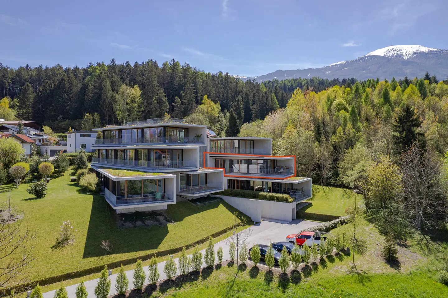 Modern multi-story home with concrete and glass design, green roofs, and mountain backdrop. Cars parked in front.