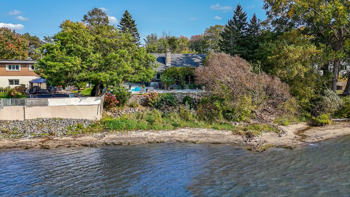 Waterfront property with a stone house, pool, and mature trees on a sunny day. The water is blue and the sky is clear.