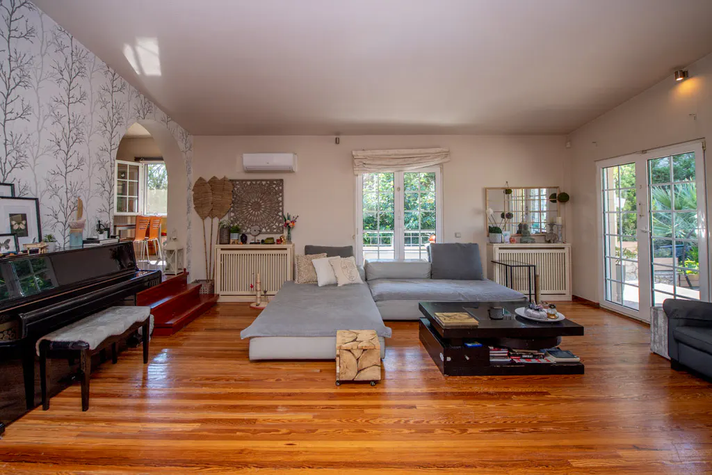 Bright living room with hardwood floors, a gray sectional sofa, and a black piano. Large windows and doors offer views of the greenery outside.