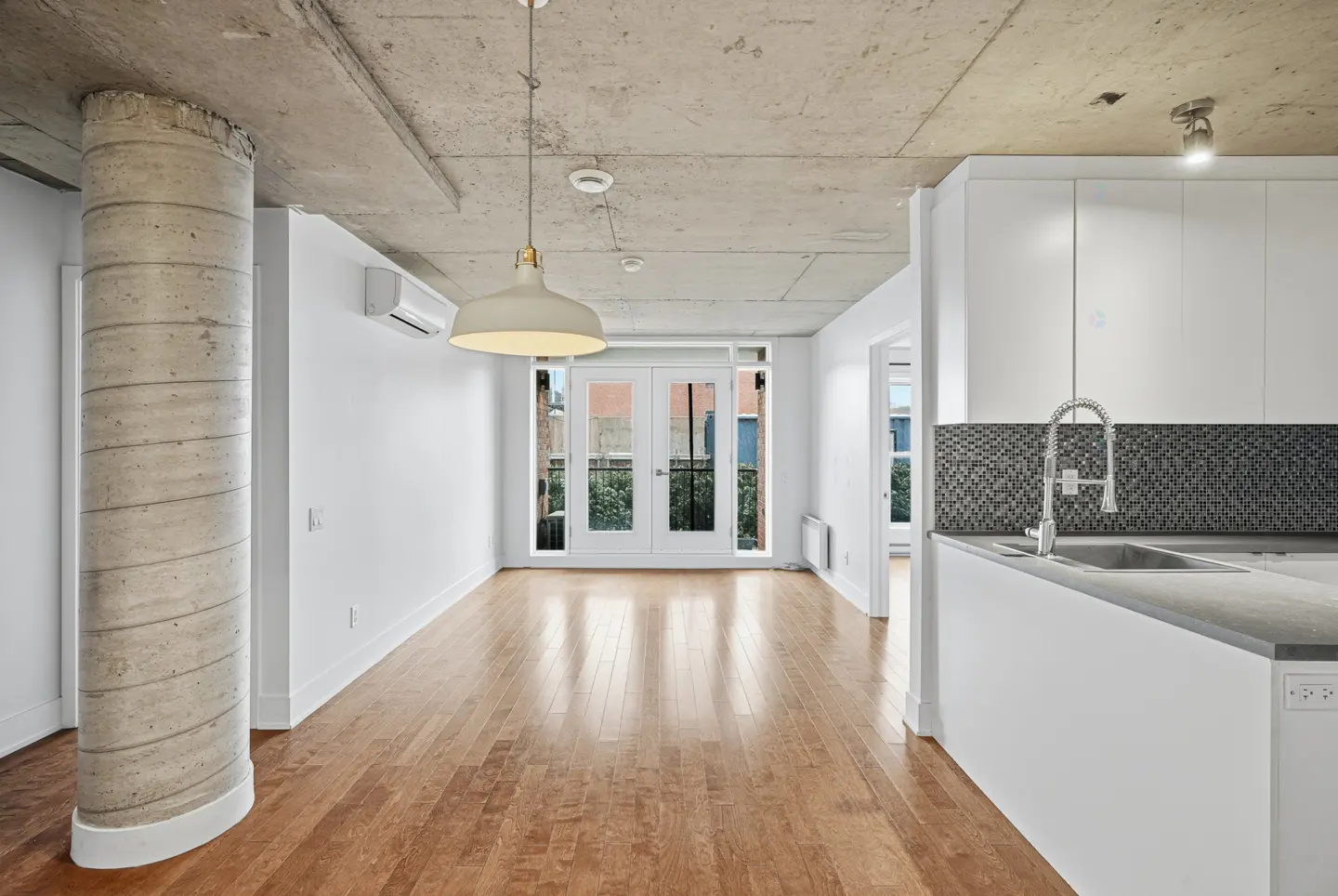 Bright, modern apartment interior with hardwood floors, white walls, and a concrete pillar. French doors lead to a balcony. Kitchen with white cabinets.