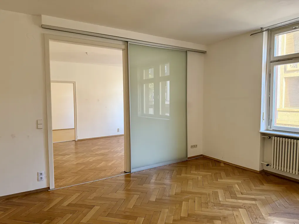 Bright room with herringbone wood floors, white walls, and a sliding glass door to another room. A window is on the right.