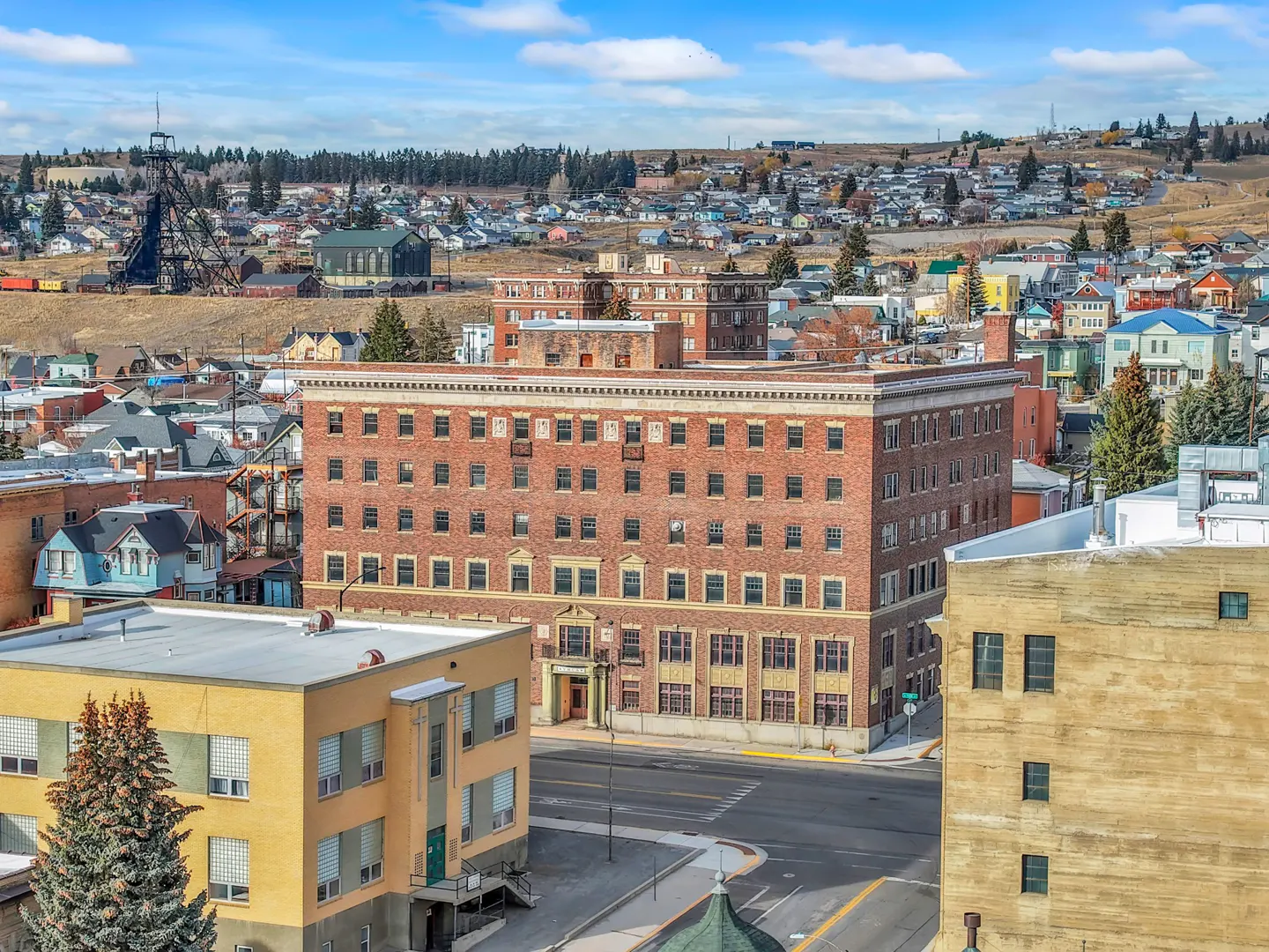Cityscape view of Butte, Montana, featuring brick buildings, a mine headframe, and houses on a hillside under a blue sky.