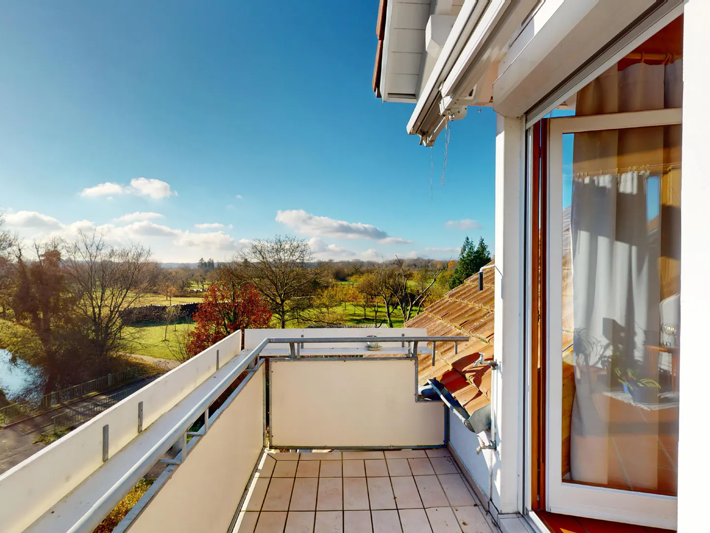Balcony view with white railings, beige tile floor, and open window. A green field and blue sky are visible in the background.