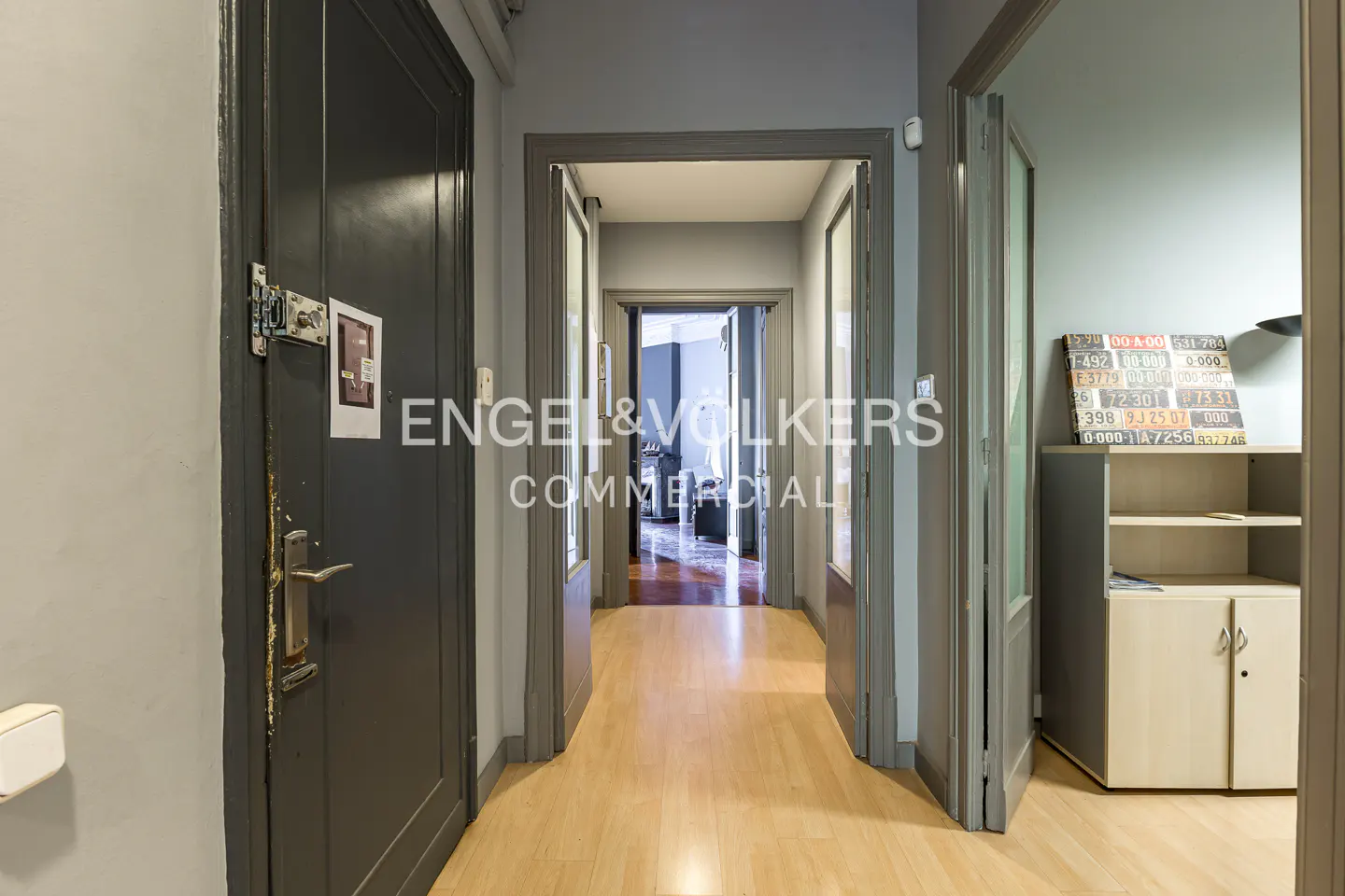 Hallway with light wood floors, gray walls, and gray doors. An open doorway leads to another room. Engel & Volkers logo is visible.