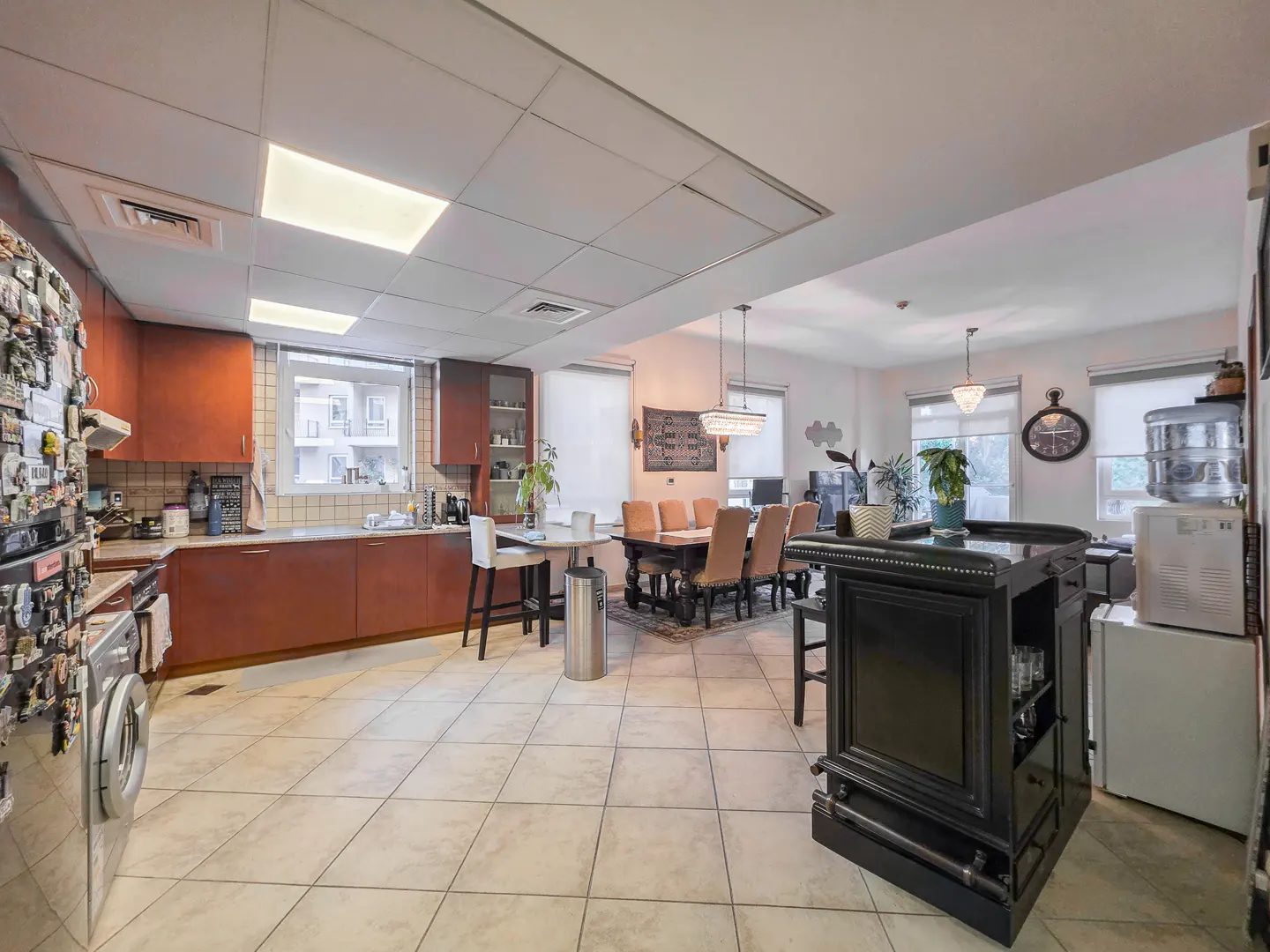 A bright, open-concept kitchen and dining area with tile flooring. Brown cabinets line the kitchen, and a black bar stands near a dining table with tan chairs.