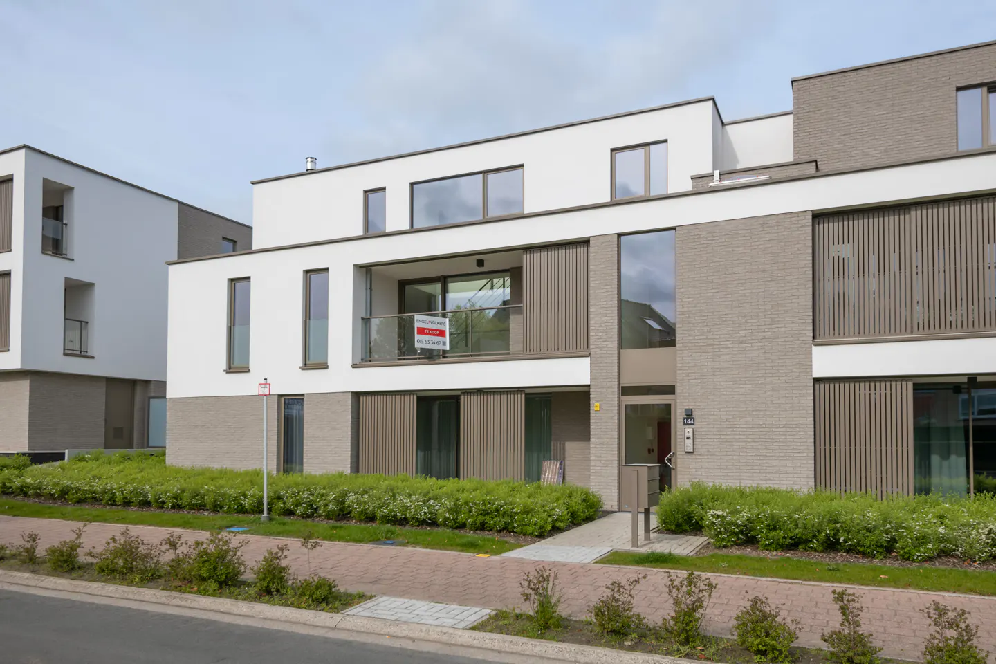 Modern apartment building with white and gray brick facade. A "For Sale" sign hangs on the balcony. Green bushes line the walkway.