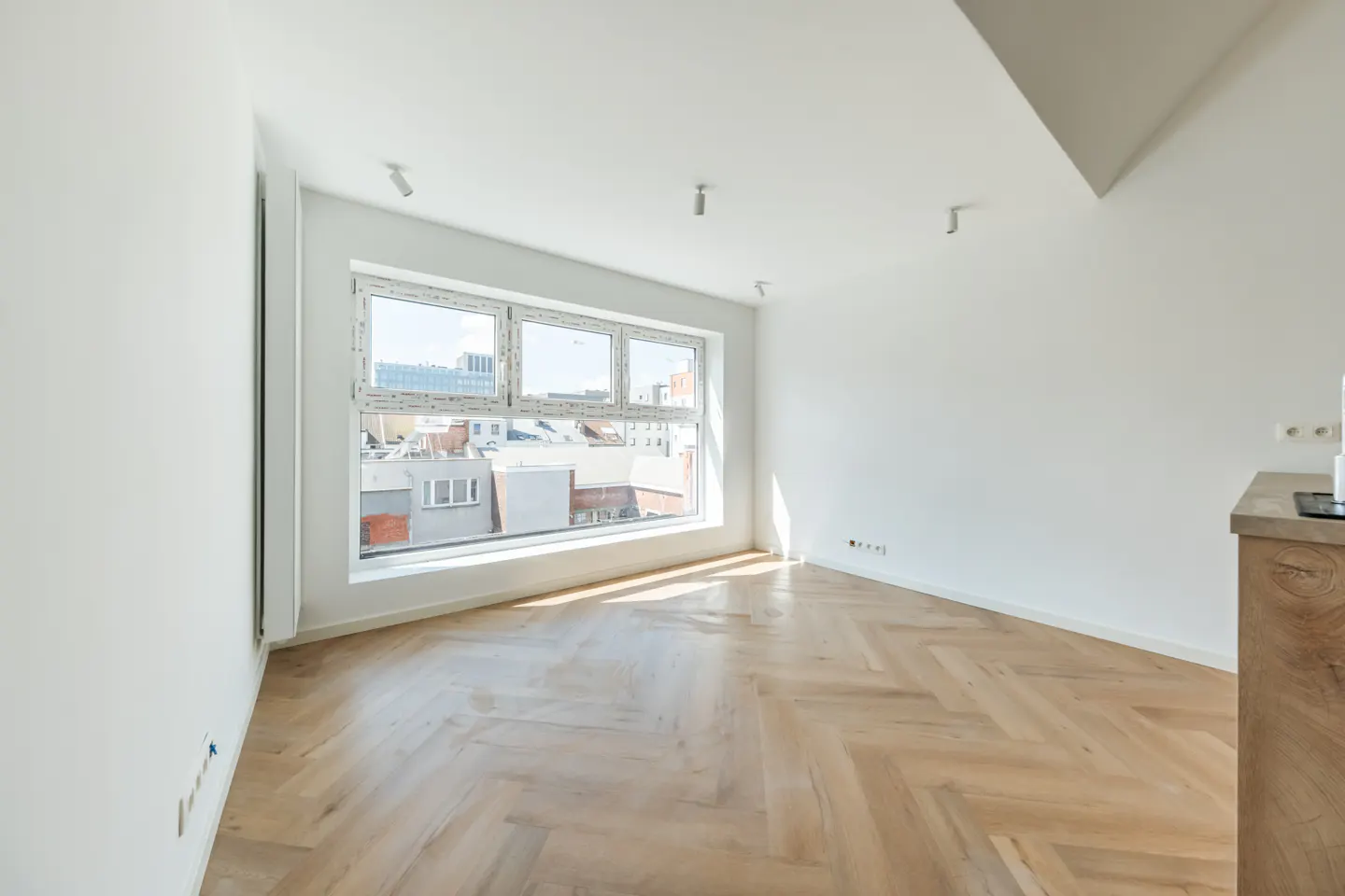 Bright, empty room with herringbone wood floors, white walls, and a large window showing city buildings.