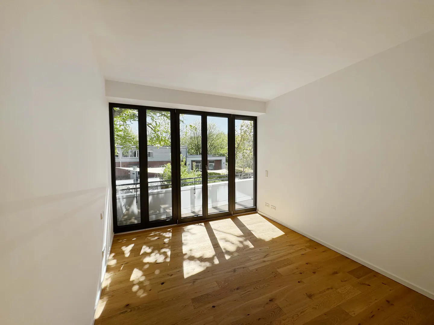 Empty room with light wood floors, white walls, and large black-framed glass doors opening to a balcony with tree views.