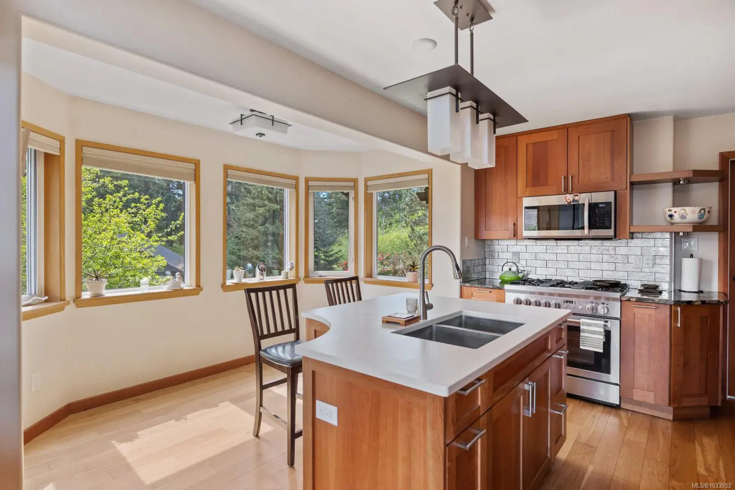 Bright kitchen with wood cabinets, white countertops, and stainless steel appliances. Large windows offer a view of lush greenery.