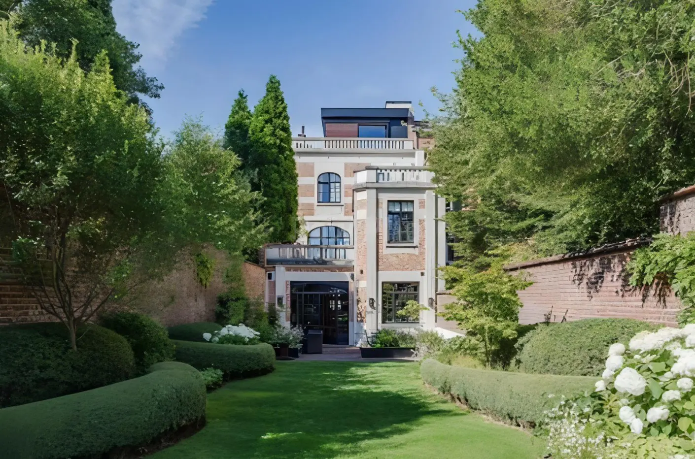 A three-story brick and stone house with a manicured lawn and hedges. Trees surround the property.