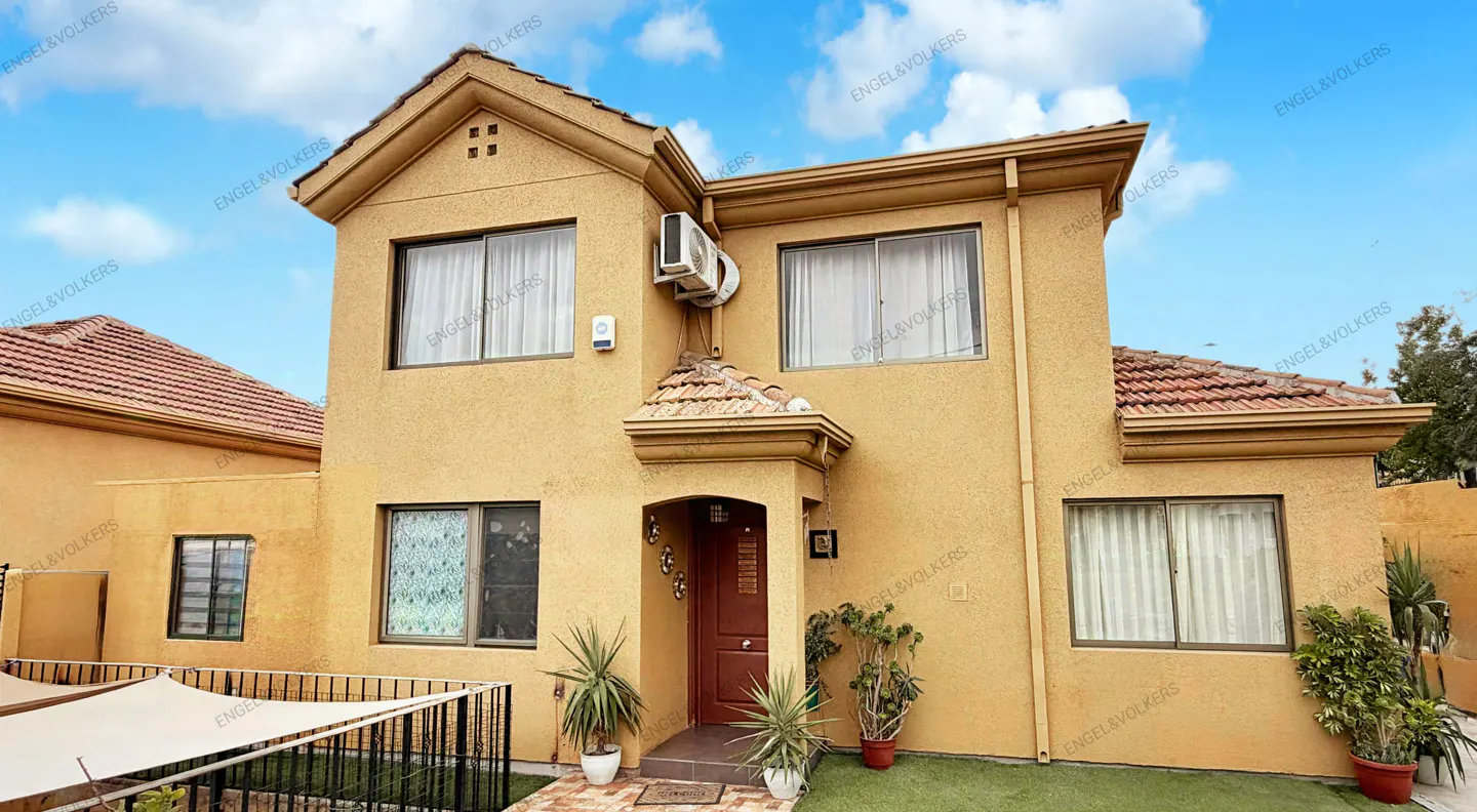 Two-story tan house with a red tile roof, brown door, and green plants in front. Blue sky with clouds above.