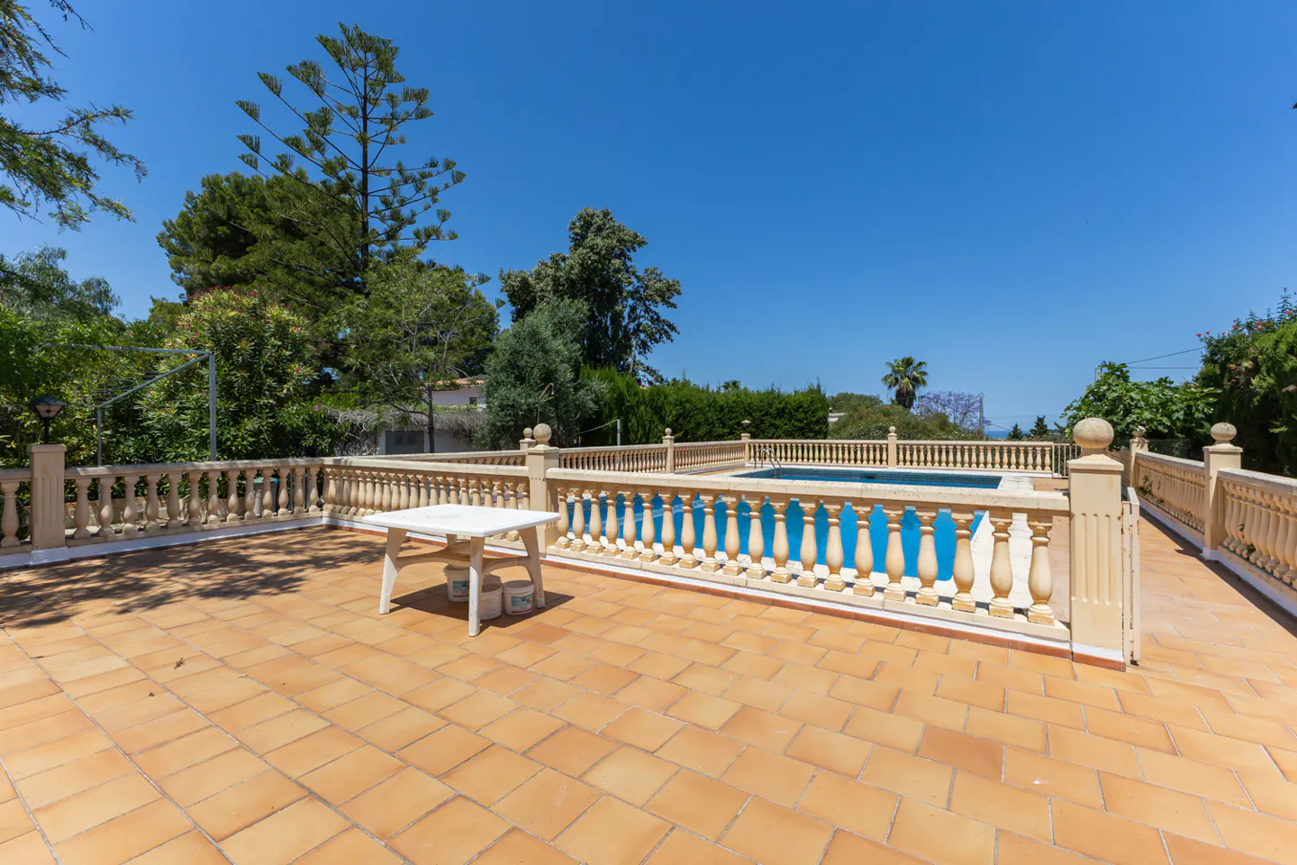 Terracotta tiled patio with a white table and a stone balustrade overlooking a blue swimming pool and lush green trees under a clear blue sky.
