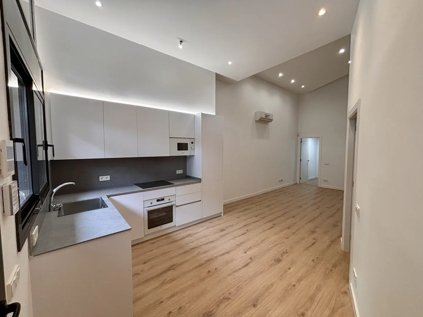 Bright, modern apartment with white cabinets, gray countertops, and wood floors. A black-framed window is on the left.