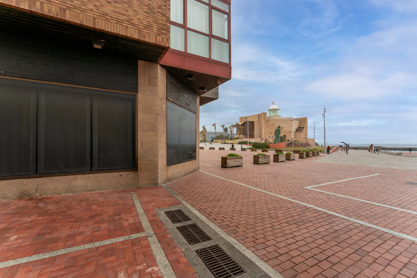 Brick building with dark windows overlooks a brick-paved plaza. In the distance, a building with a green dome sits near the ocean.
