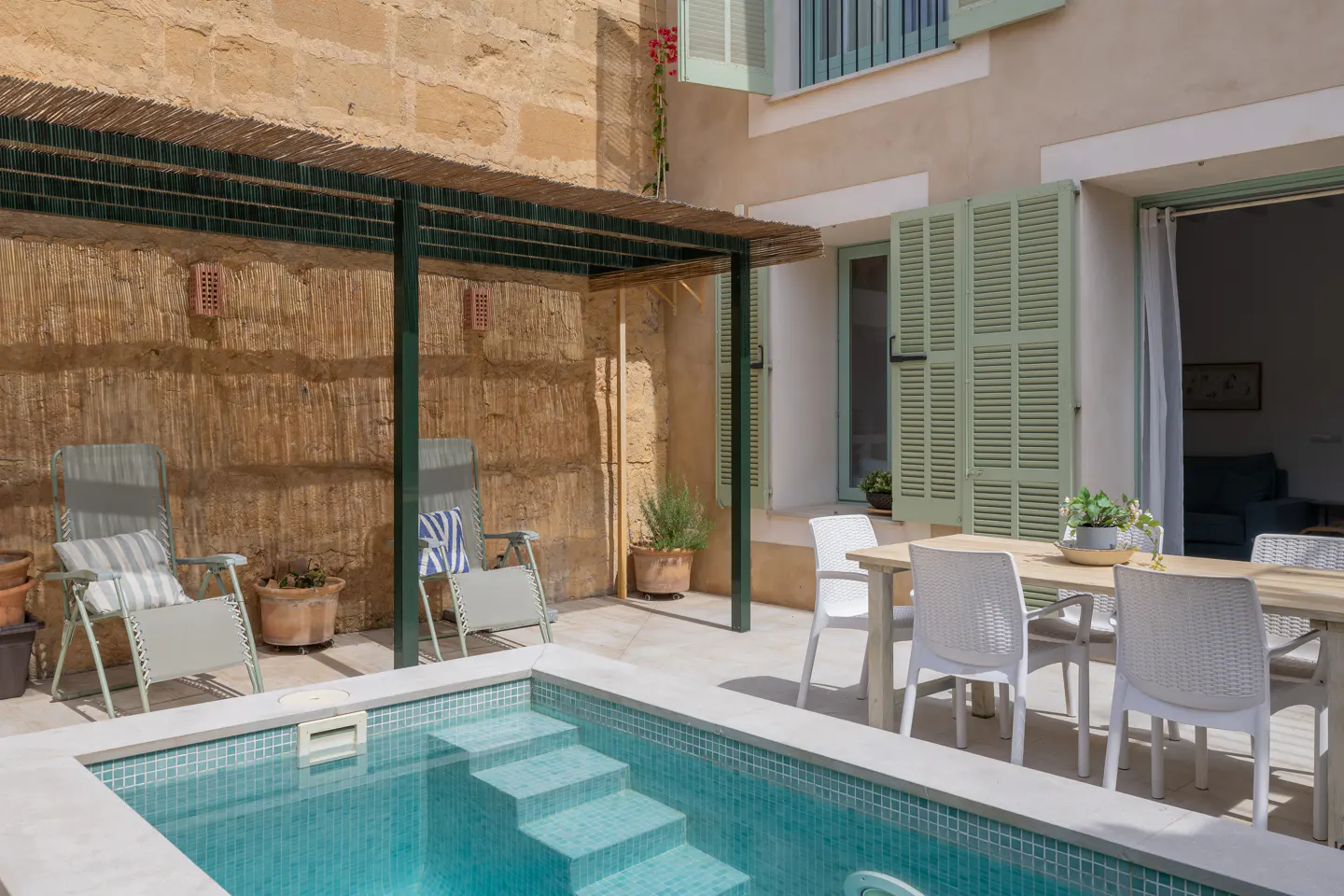 Outdoor patio with a small pool, chairs, and a dining table. The building has light green shutters and a stone wall.