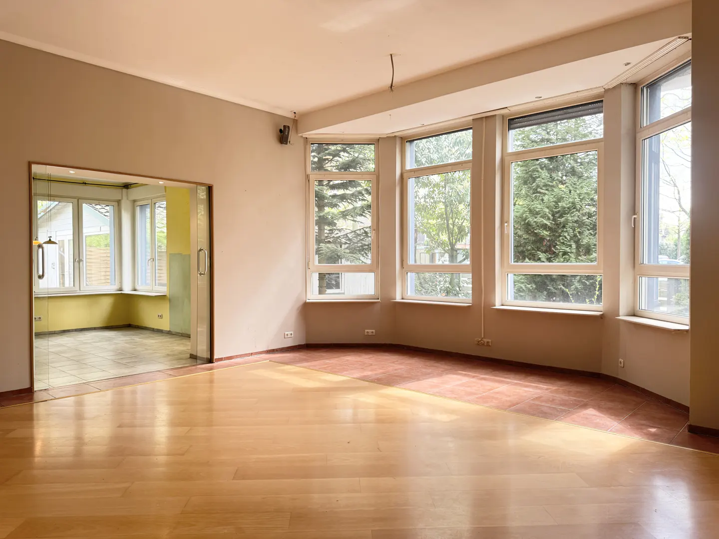 Empty room with light wood floors, beige walls, and large windows showing trees. A doorway leads to another room with yellow walls.