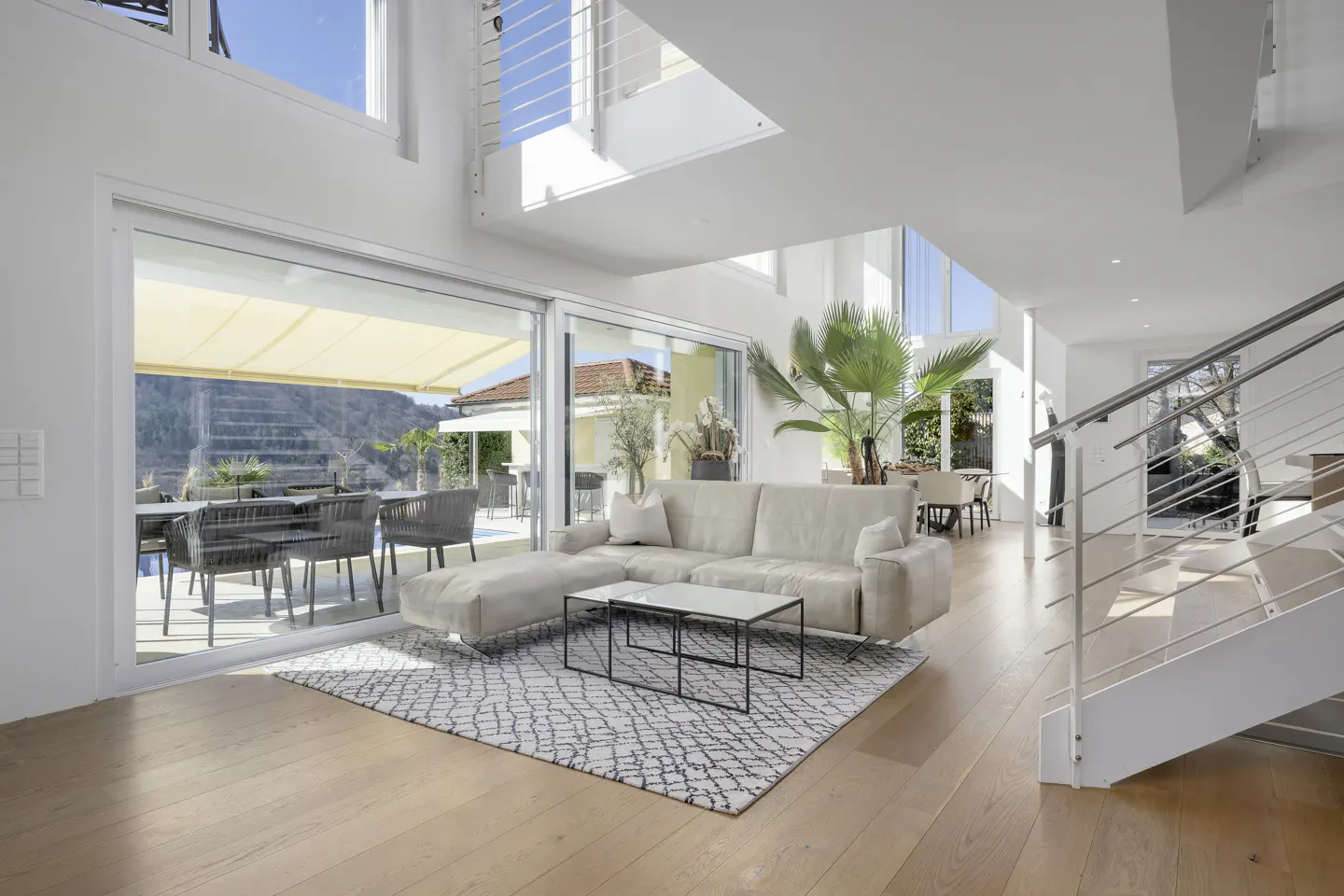 Bright, modern living room with a beige sectional sofa, black coffee tables, and a patterned rug on a wood floor. Sliding glass doors open to a patio with outdoor seating and a pool. White walls and staircase.