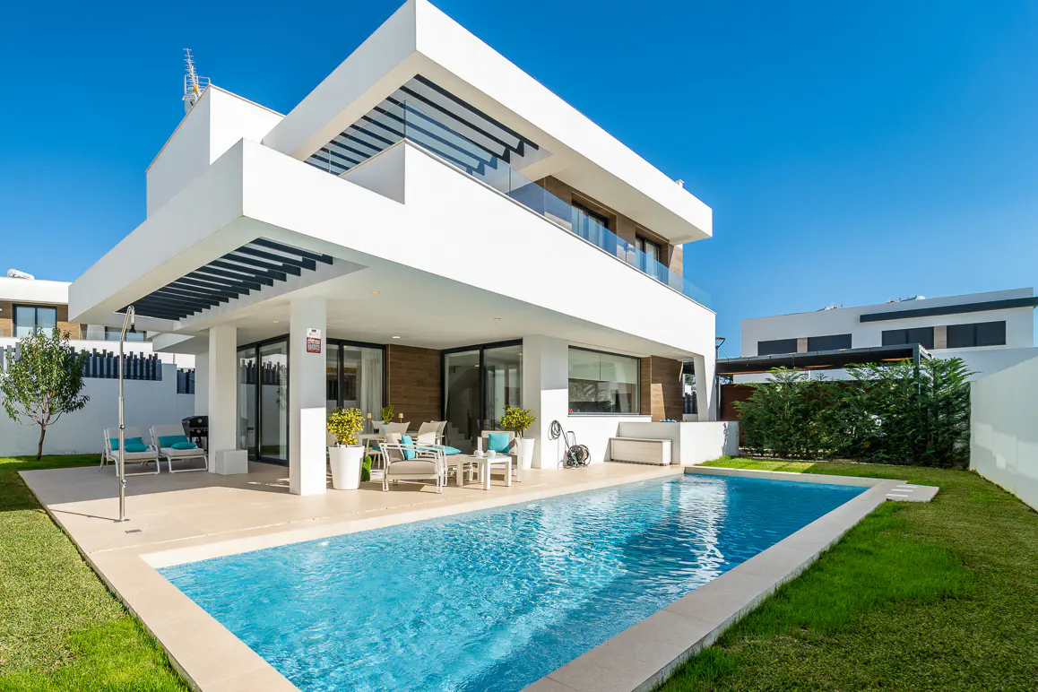 Modern white two-story house with a blue swimming pool, patio furniture, and green grass under a clear blue sky.