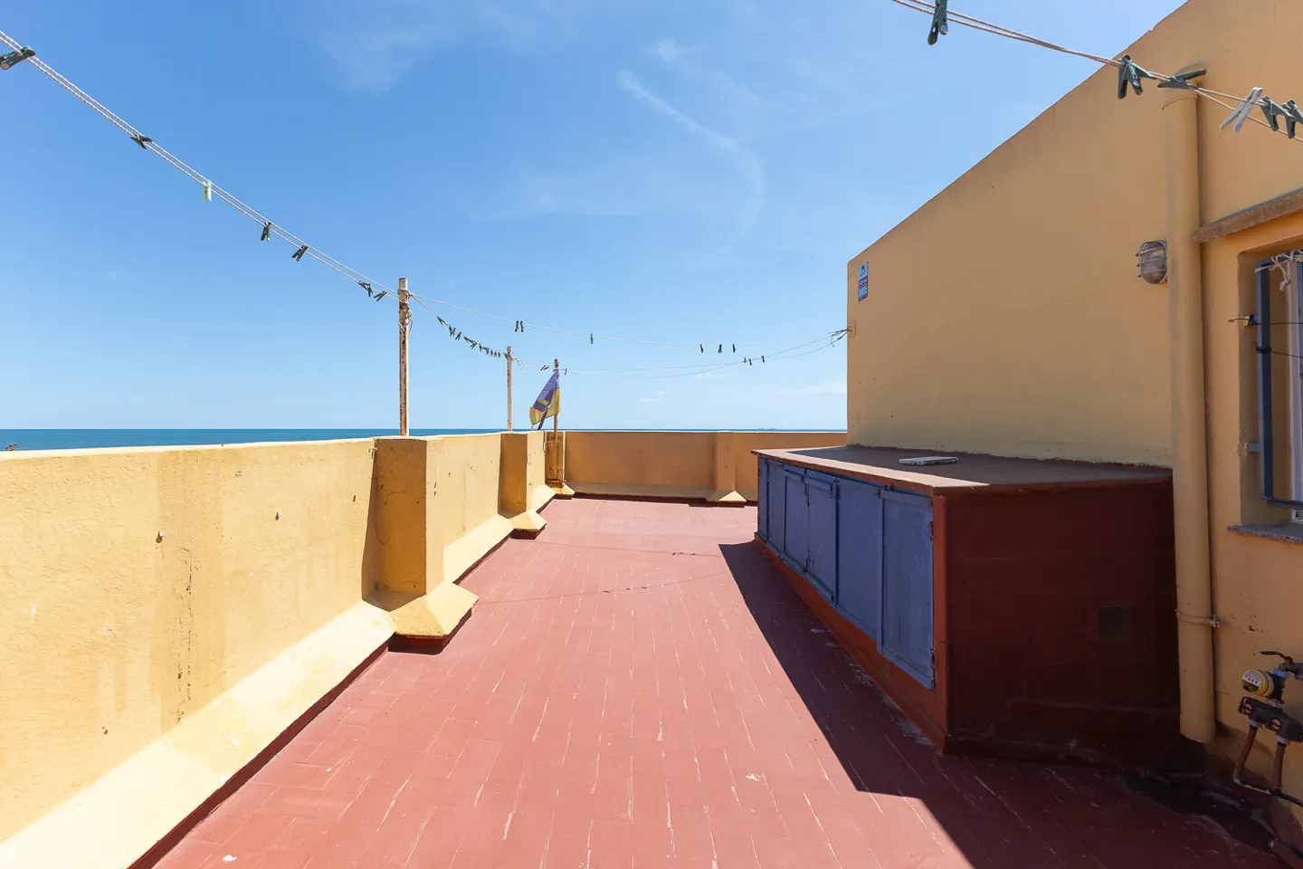 Rooftop view with red brick floor, yellow walls, and blue storage. Clothesline with clothespins stretches across the blue sky.
