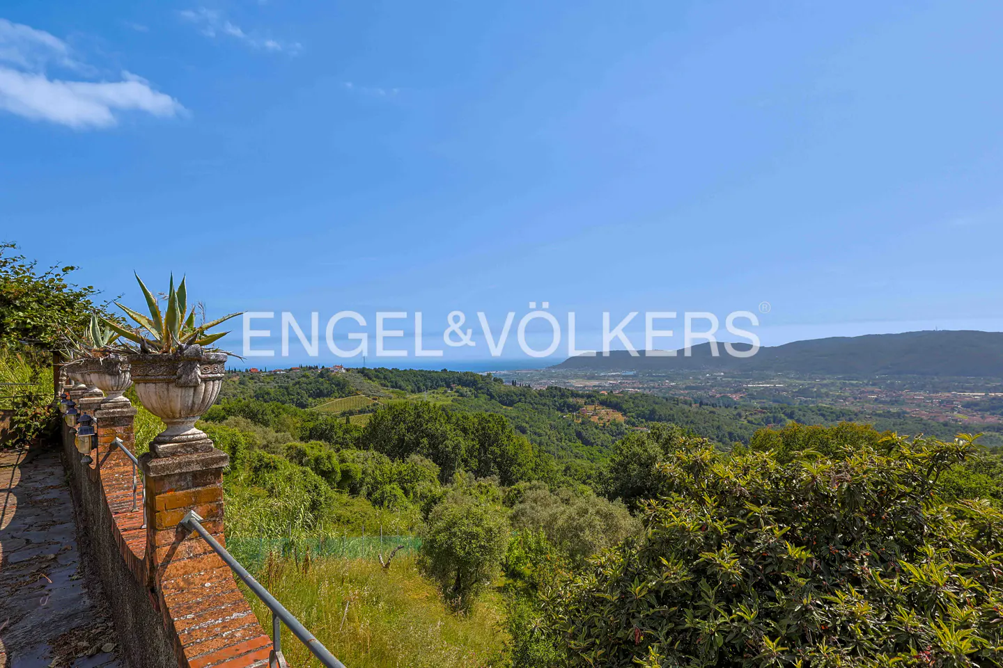 A stone terrace with potted agave plants overlooks a lush green valley, rolling hills, and a distant blue sea under a sky.