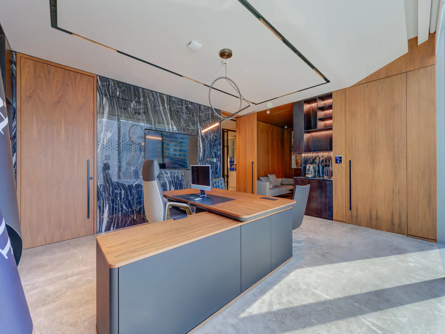 A modern office with a wood and gray desk, computer, and chair. Marble wall and wood cabinets add texture. A gray sofa is visible in the background.