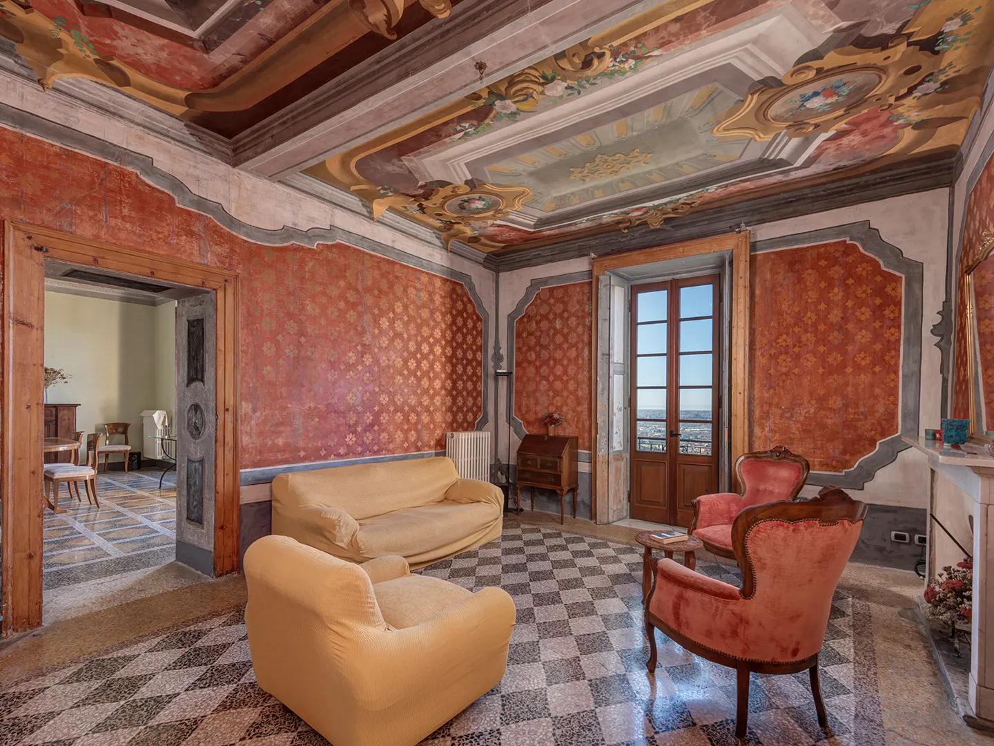 Ornate living room with painted ceiling, red patterned walls, and checkered floor. Yellow sofa and red chairs furnish the space.