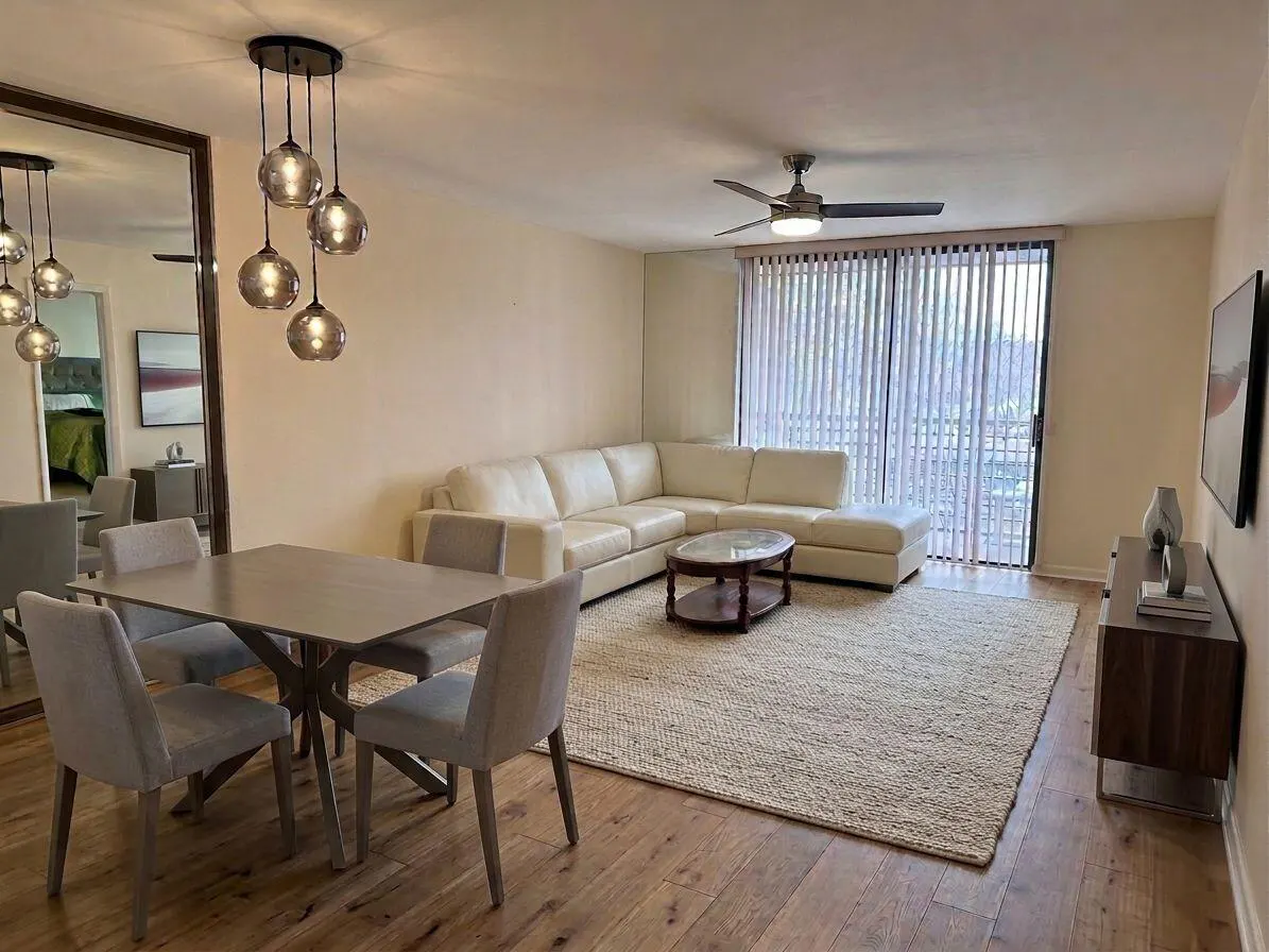 Living room with dining area features a white sectional sofa, wood floors, and a dining table with four chairs. A ceiling fan and sliding glass doors are visible.