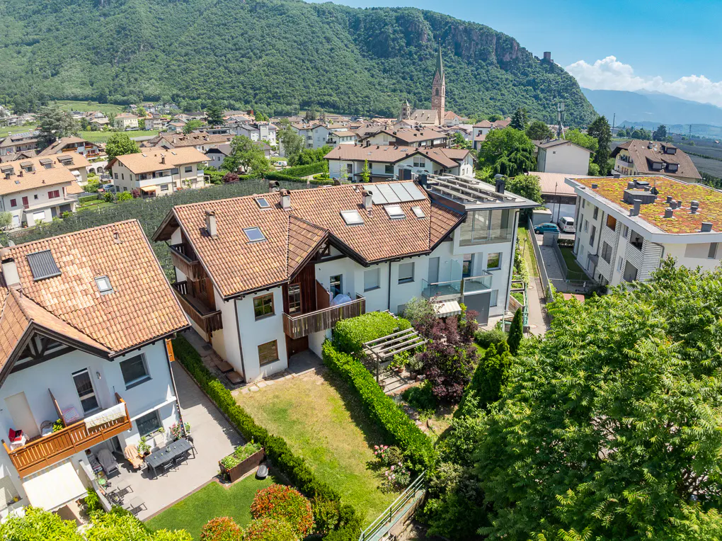 Aerial view of white residential buildings with terracotta roofs, green lawns, and a mountain village backdrop.