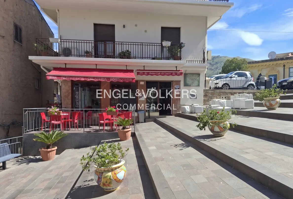 Exterior of a two-story white building with a red awning and outdoor seating. Potted plants decorate the steps.