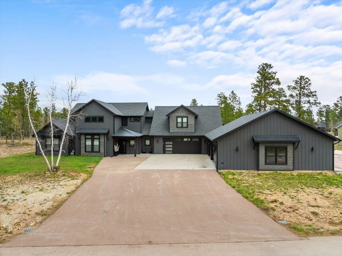 Modern dark gray farmhouse with a large concrete driveway, surrounded by trees under a blue sky with light clouds.