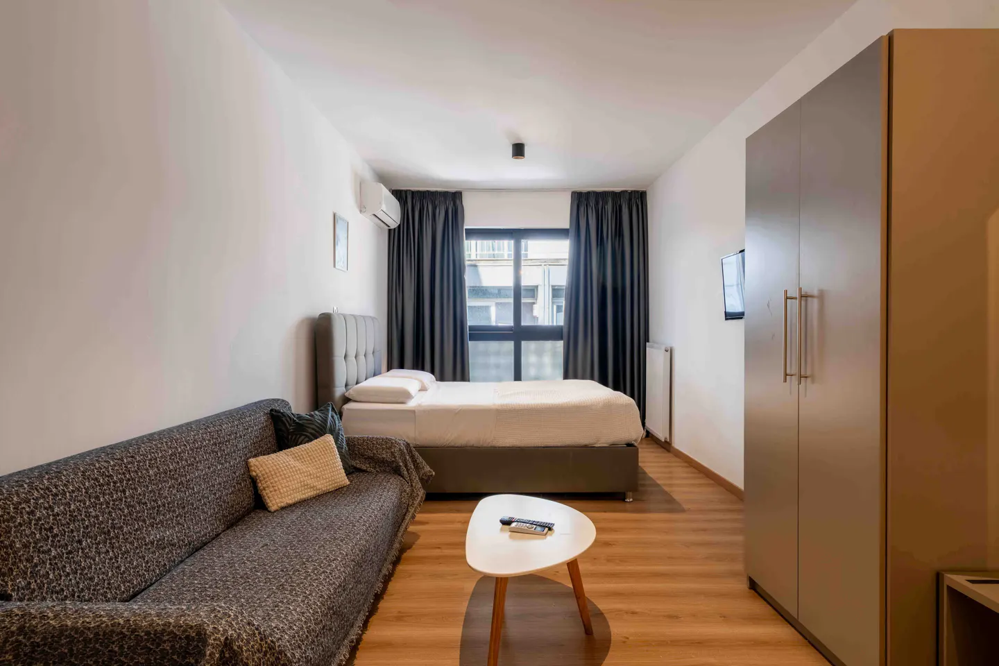 Studio apartment with a gray sofa, bed, and wardrobe. A small white table sits on the wooden floor. Curtains frame the window.