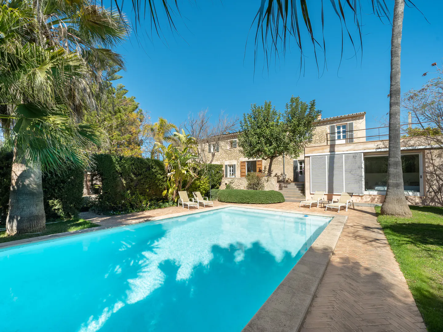A sunny backyard with a turquoise pool, lounge chairs, and a stone house with trees.