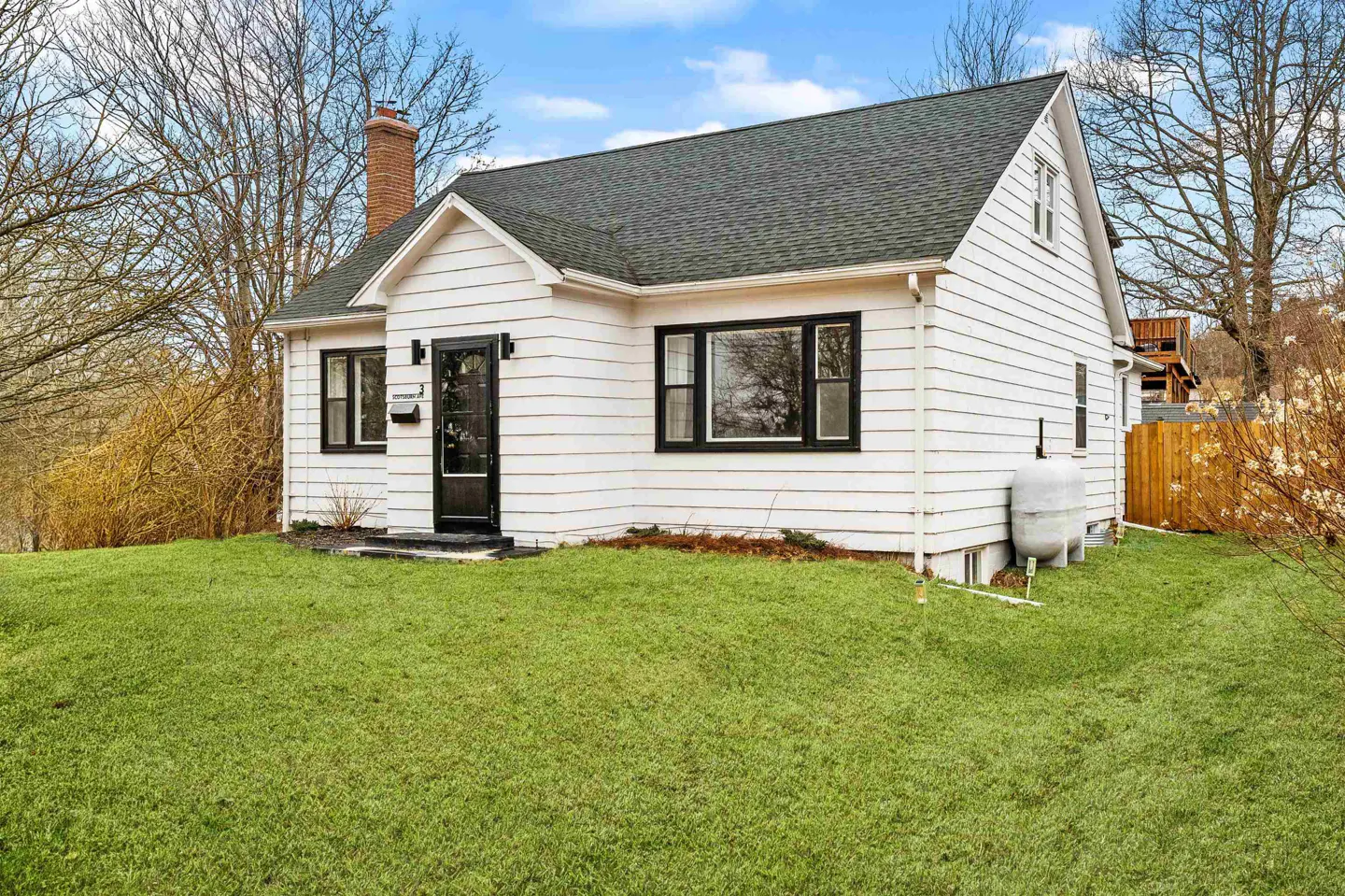 Exterior view of a white, one-story house with a green roof, black trim, and a brick chimney. A green lawn is in the foreground.