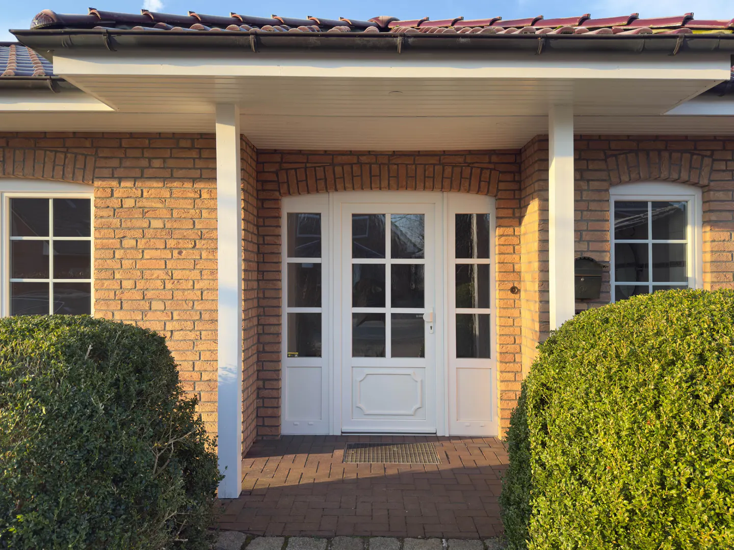 Front entrance of a brick house with a white door and windows. Green bushes flank the walkway.